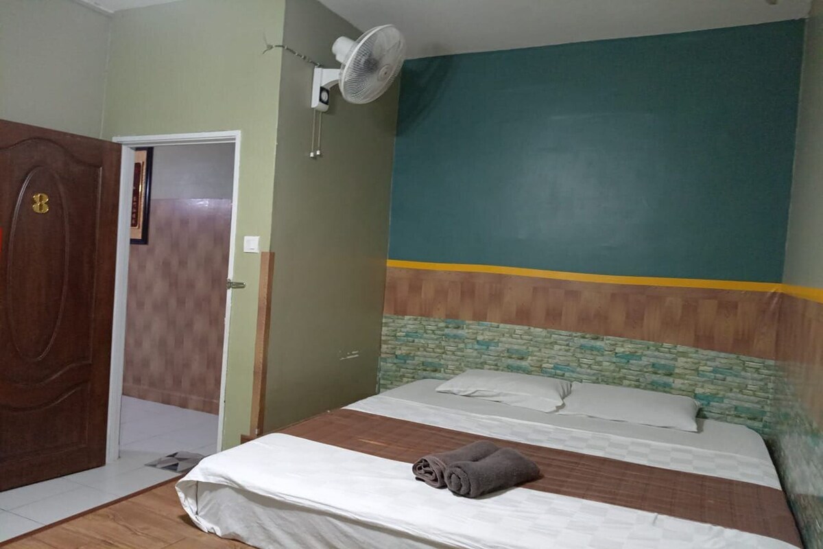 A bedroom with a bed, a wooden door, a wall-mounted fan, and a green and yellow painted wall.