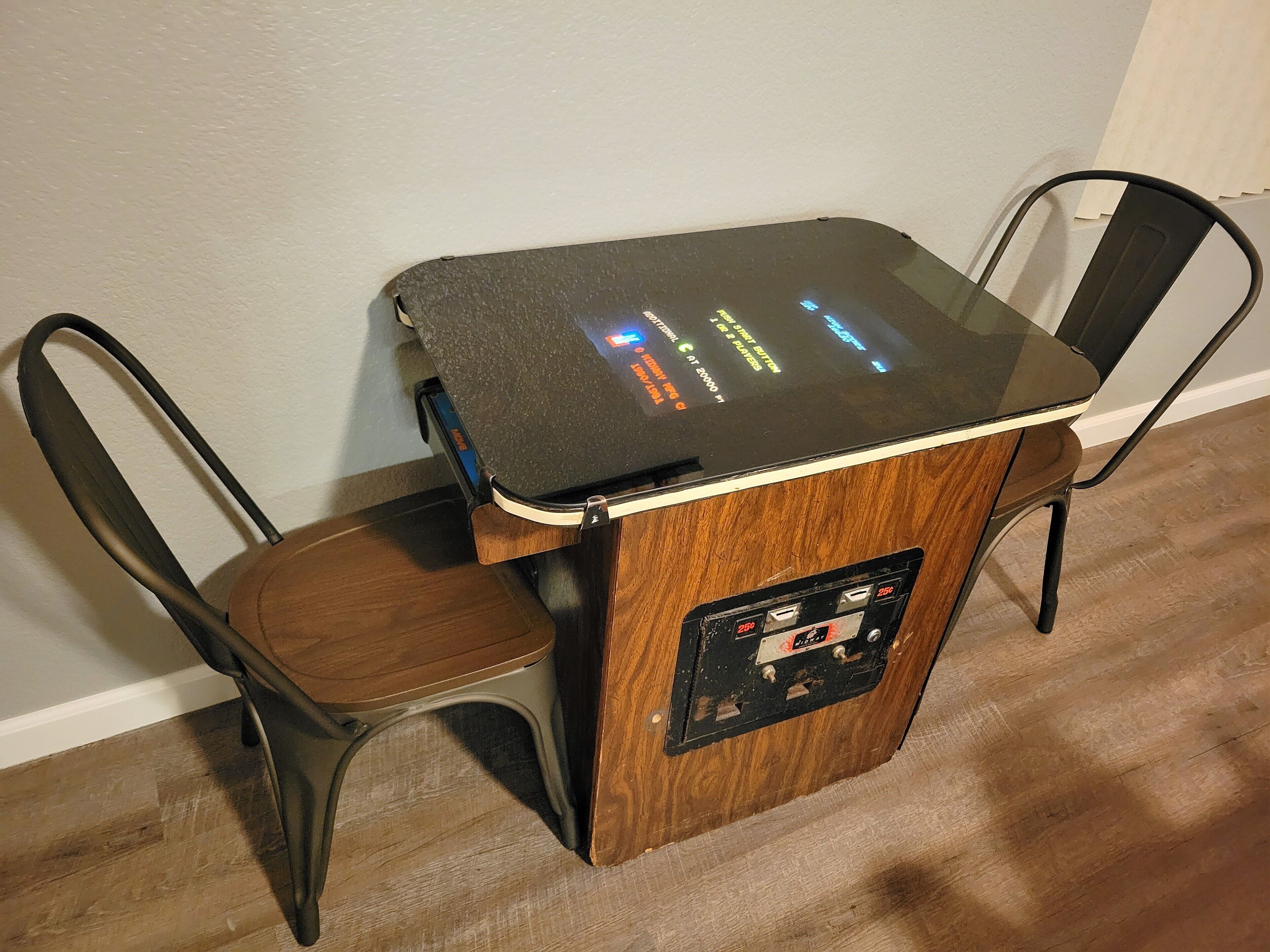 A small wooden table with a glass top and a control panel, accompanied by two metal chairs.