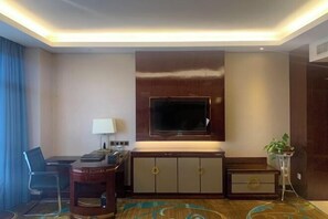A hotel room with a large flat-screen TV mounted on the wall, a desk with a chair, a small side table, and a potted plant. - Xixia International Hotel (Cheng County)