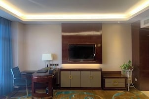 Habitación de hotel con una televisión de pantalla plana grande montada en la pared, un escritorio con una silla, una mesita auxiliar pequeña y una planta en maceta.