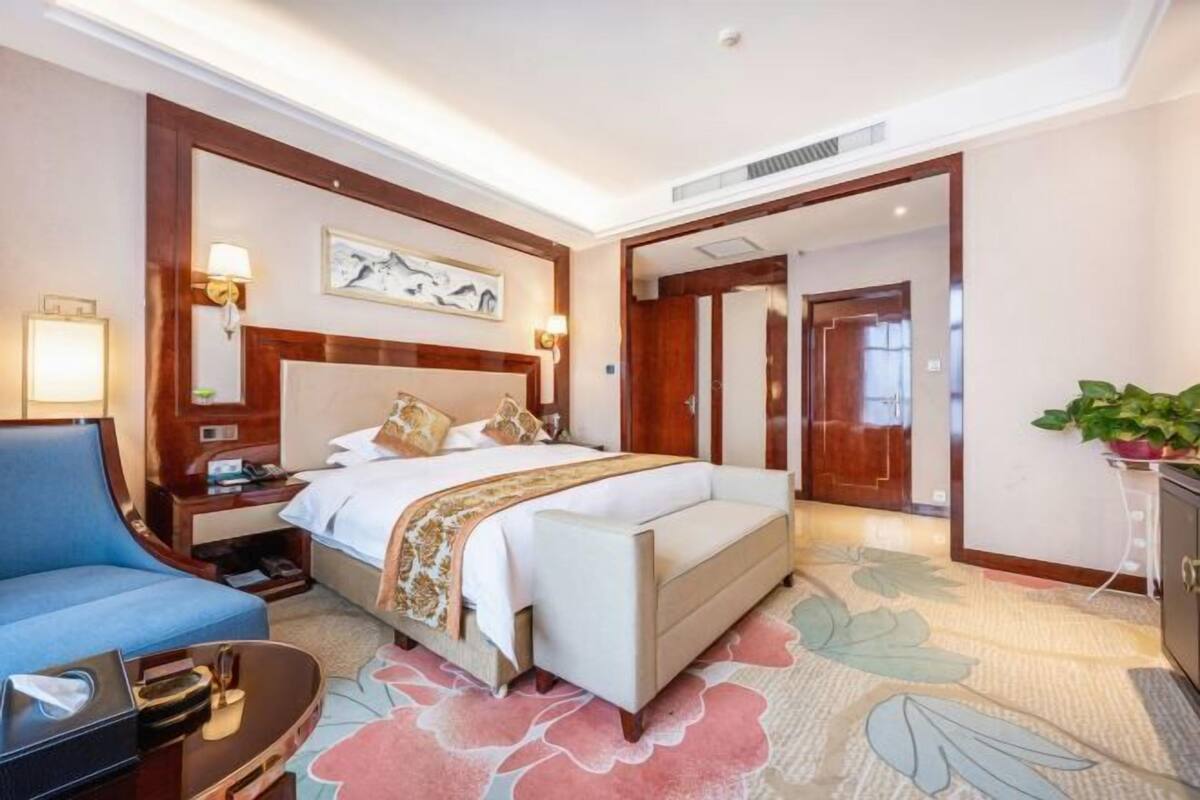 A hotel room with a bed, a blue armchair, a nightstand, a lamp, and a plant.