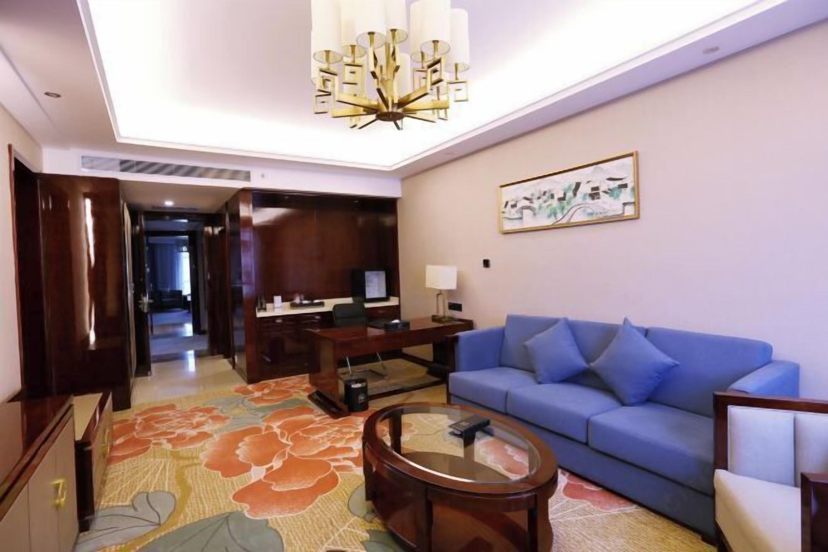 Habitación de hotel con sofá azul, una mesita de centro redonda, un escritorio con computadora y una alfombra grande con estampado floral.