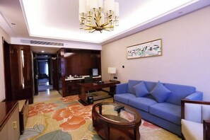 A hotel room with a blue sofa, a round coffee table, a desk with a computer, and a large floral carpet. - Xixia International Hotel (Cheng County)