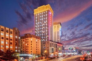 Un paisaje urbano con edificios altos, uno con señalización iluminada, y una calle concurrida con vehículos en movimiento.
