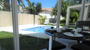 A modern outdoor dining area with a glass table and chairs, overlooking a swimming pool and a garden with palm trees.