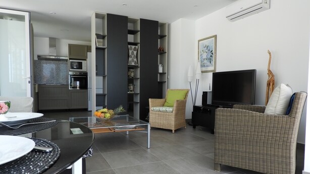 A modern living room with a dining area, a kitchen in the background, and a large flat-screen TV.