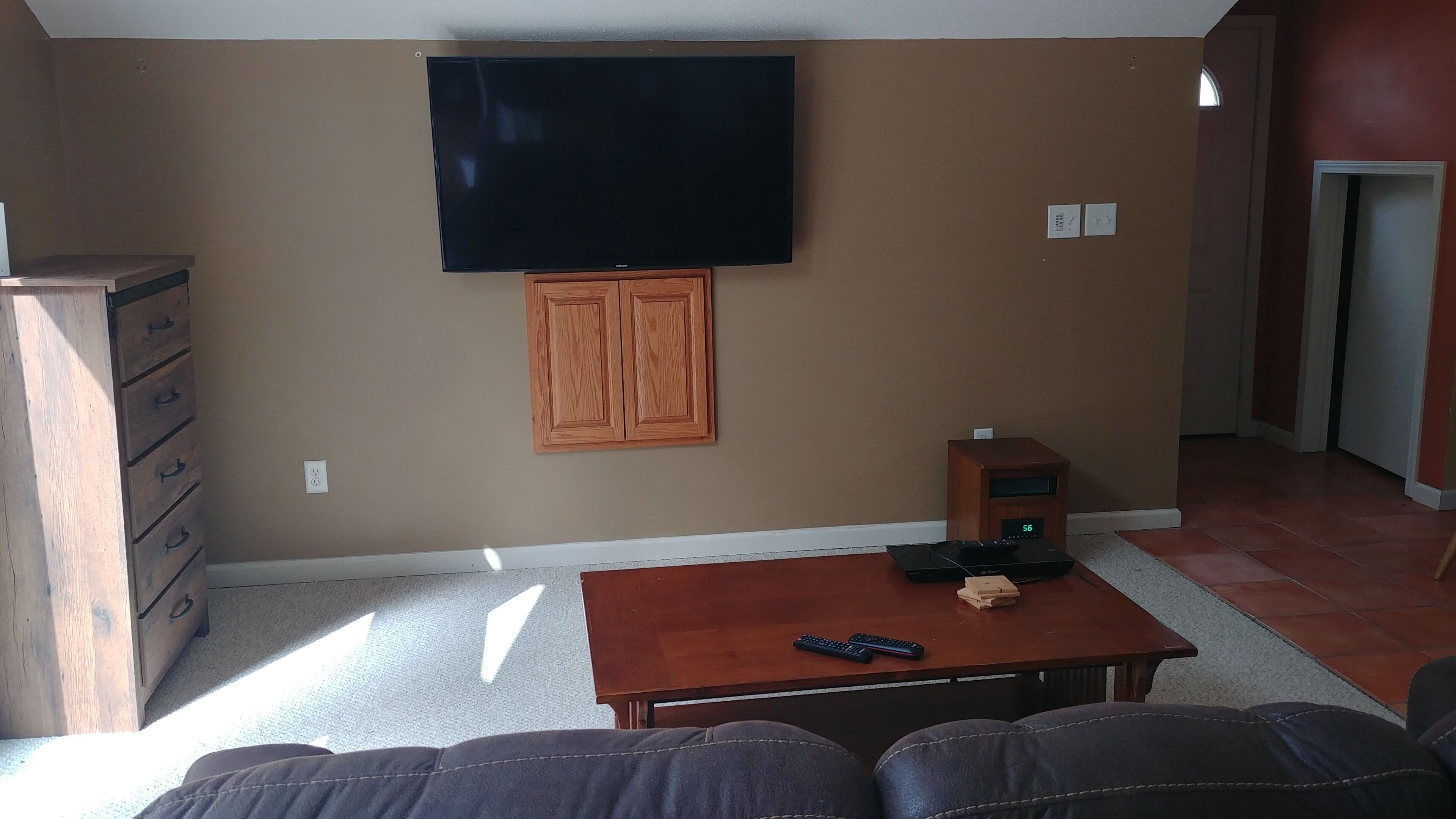 A living room with a flat-screen TV mounted on the wall, a wooden cabinet, a coffee table with a remote control, and a small speaker.