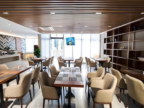 A modern dining area with multiple tables and chairs, a large screen for presentations, and a bookshelf with decorative items.