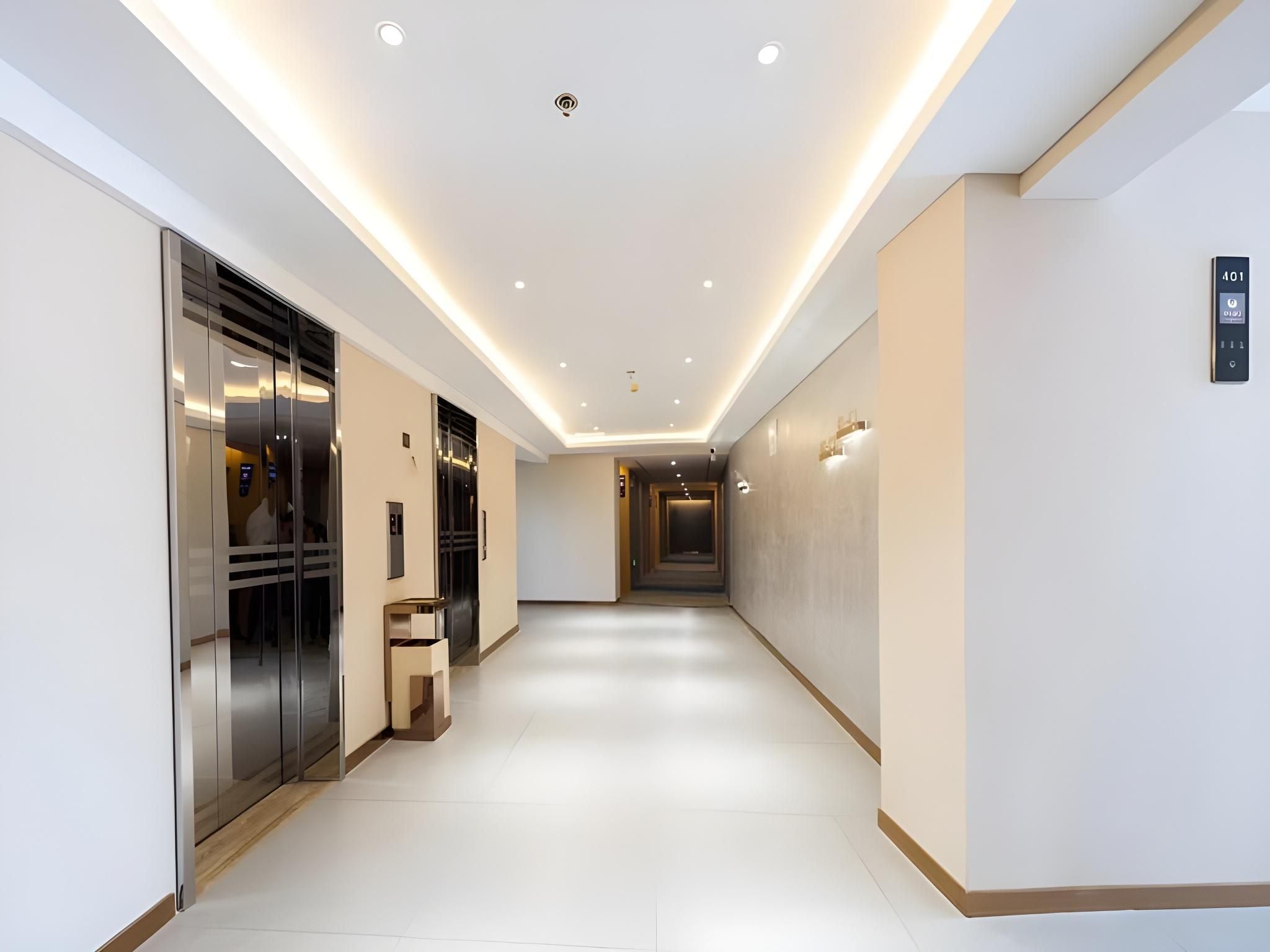 A modern hotel corridor with a glass elevator, a reception desk, and a wall-mounted elevator control panel.