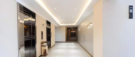 A modern hotel corridor with a glass elevator, a reception desk, and a wall-mounted elevator control panel.
