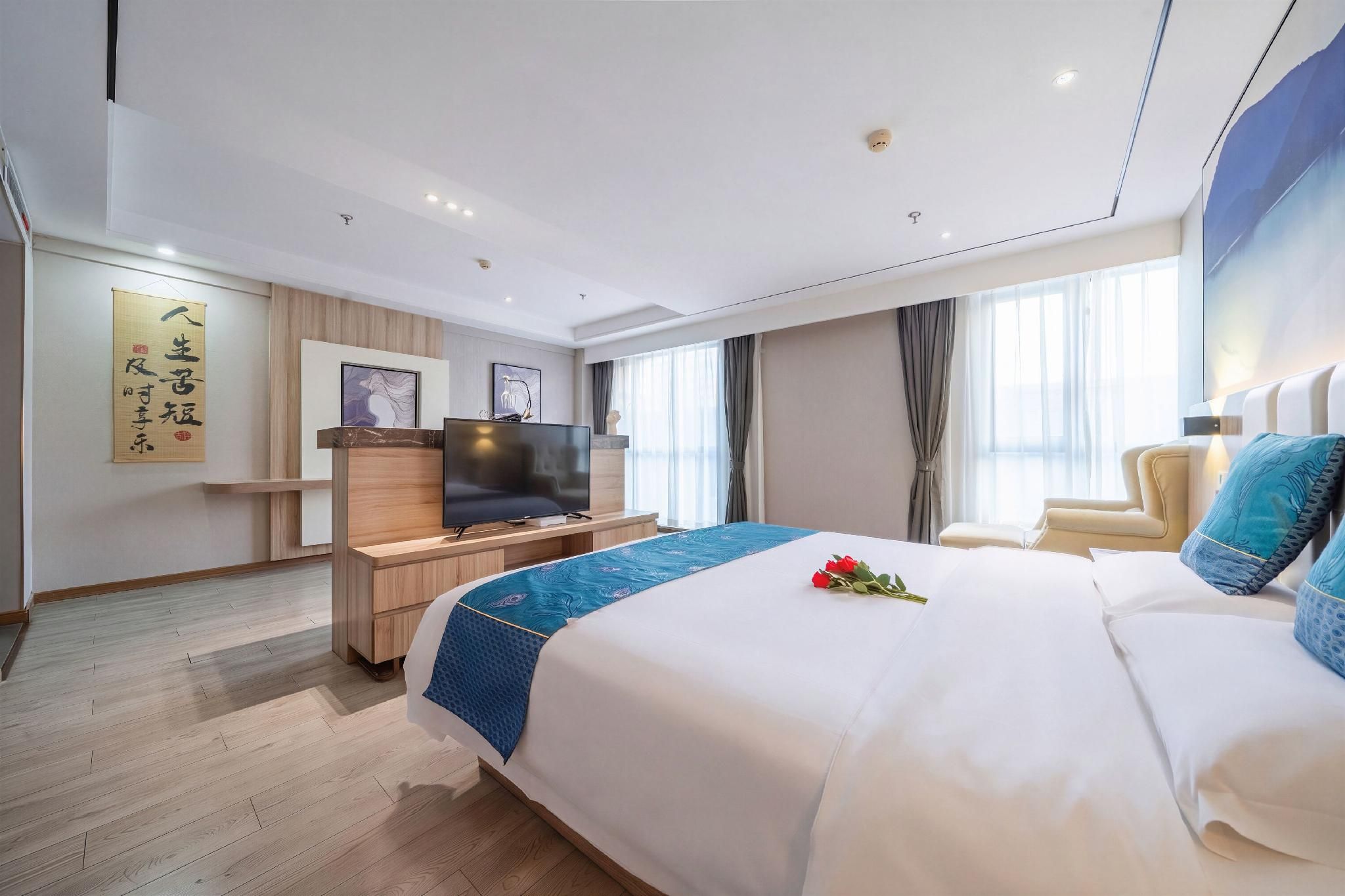 A hotel room with a large bed, a desk with a flat-screen TV, a wooden dresser, and a large window with curtains.