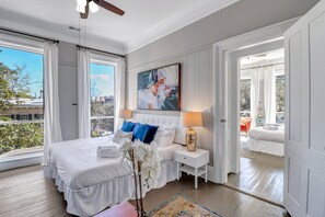 A bedroom with a large bed, white bedding, a nightstand with a lamp, a painting on the wall, and a view of the outdoors through a window.