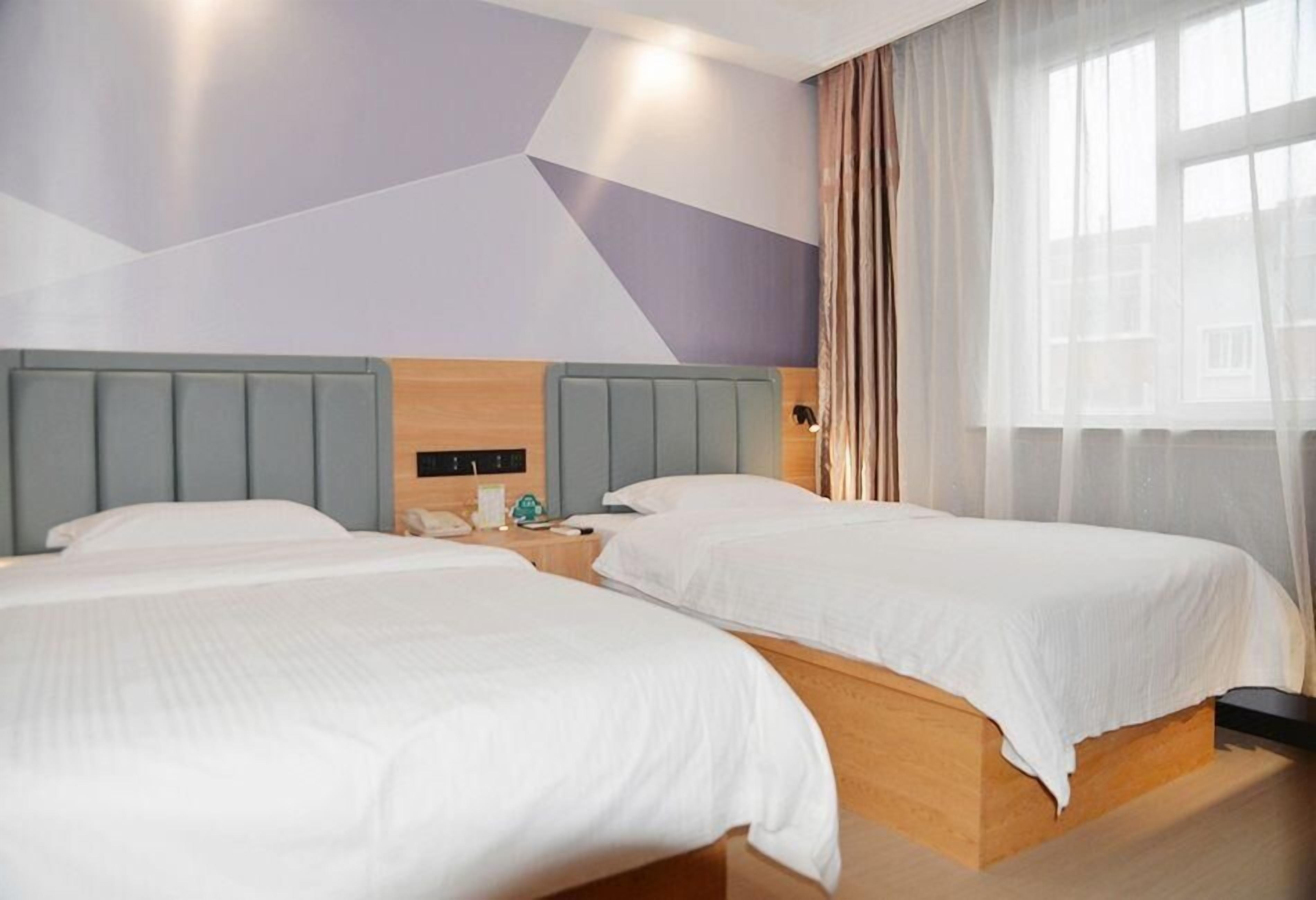 Une chambre d’hôtel avec deux lits, une tête de lit en bois, une table de chevet et une fenêtre avec des rideaux légers.