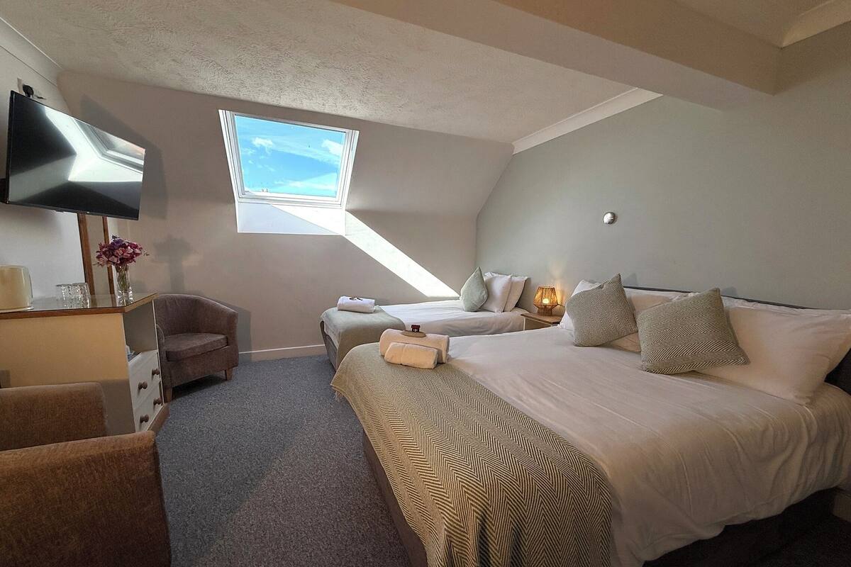 A bedroom with a large bed, a desk with a flat-screen TV, a chair, a small table, and a window with a view of the sky.