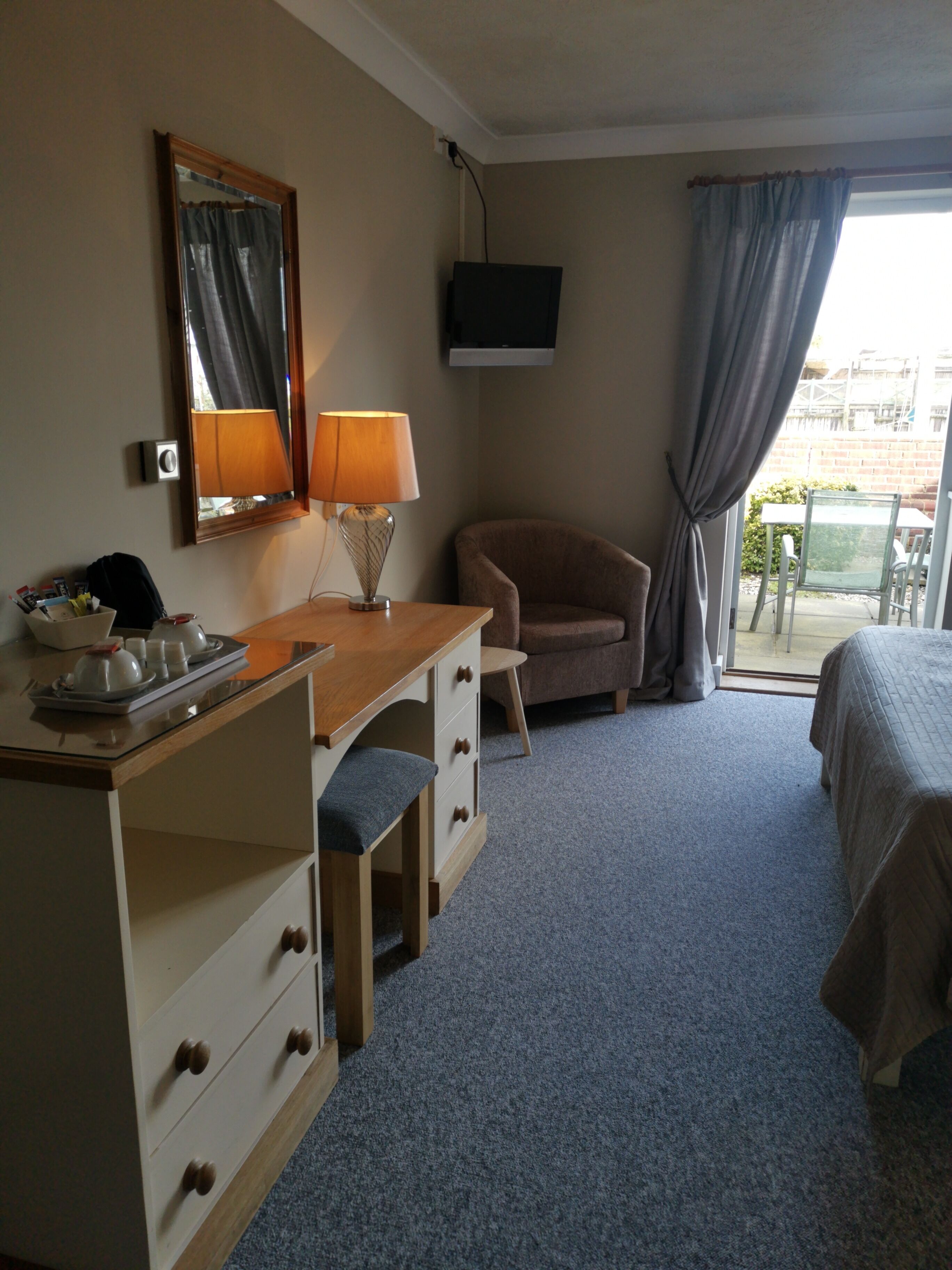 A hotel room with a wooden desk, a chair, a bed, a TV mounted on the wall, and a window with curtains.