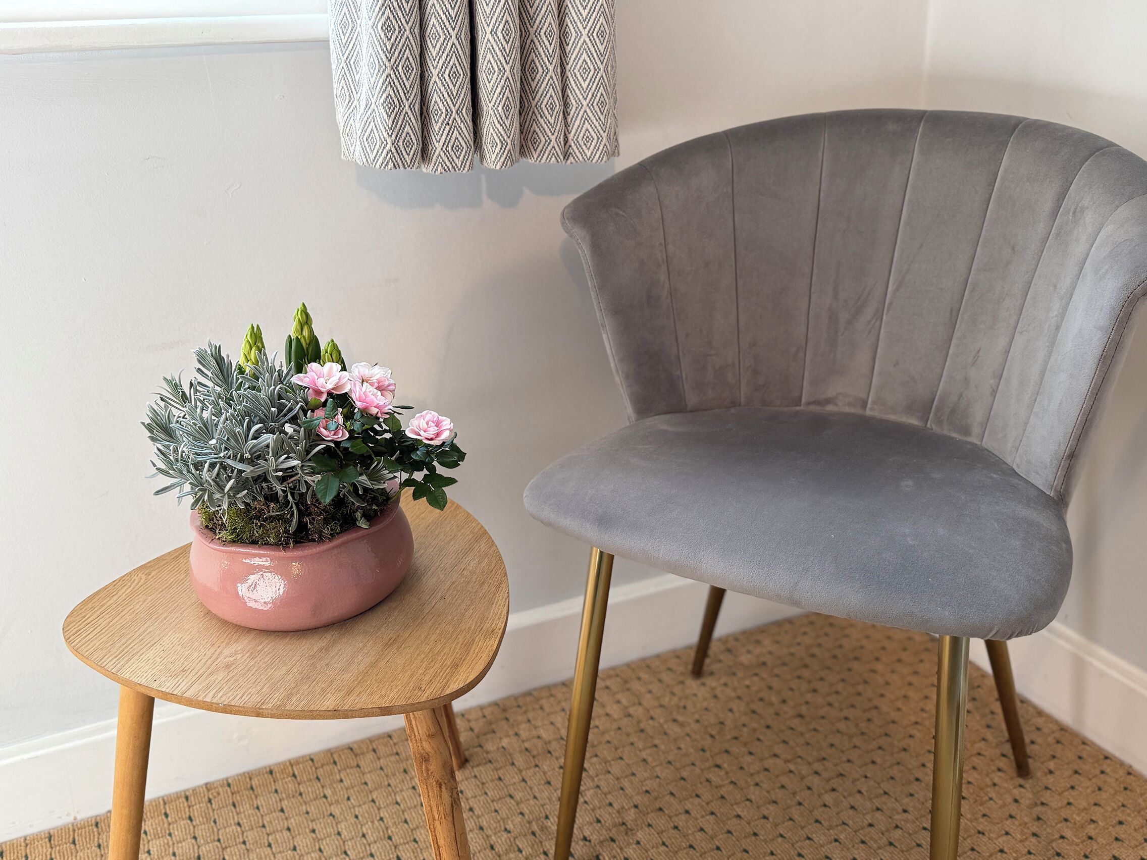 A grey upholstered chair with gold legs, a small round wooden table, and a pink potted plant with flowers and green foliage.