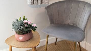 A grey upholstered chair with gold legs, a small round wooden table, and a pink potted plant with flowers and green foliage.