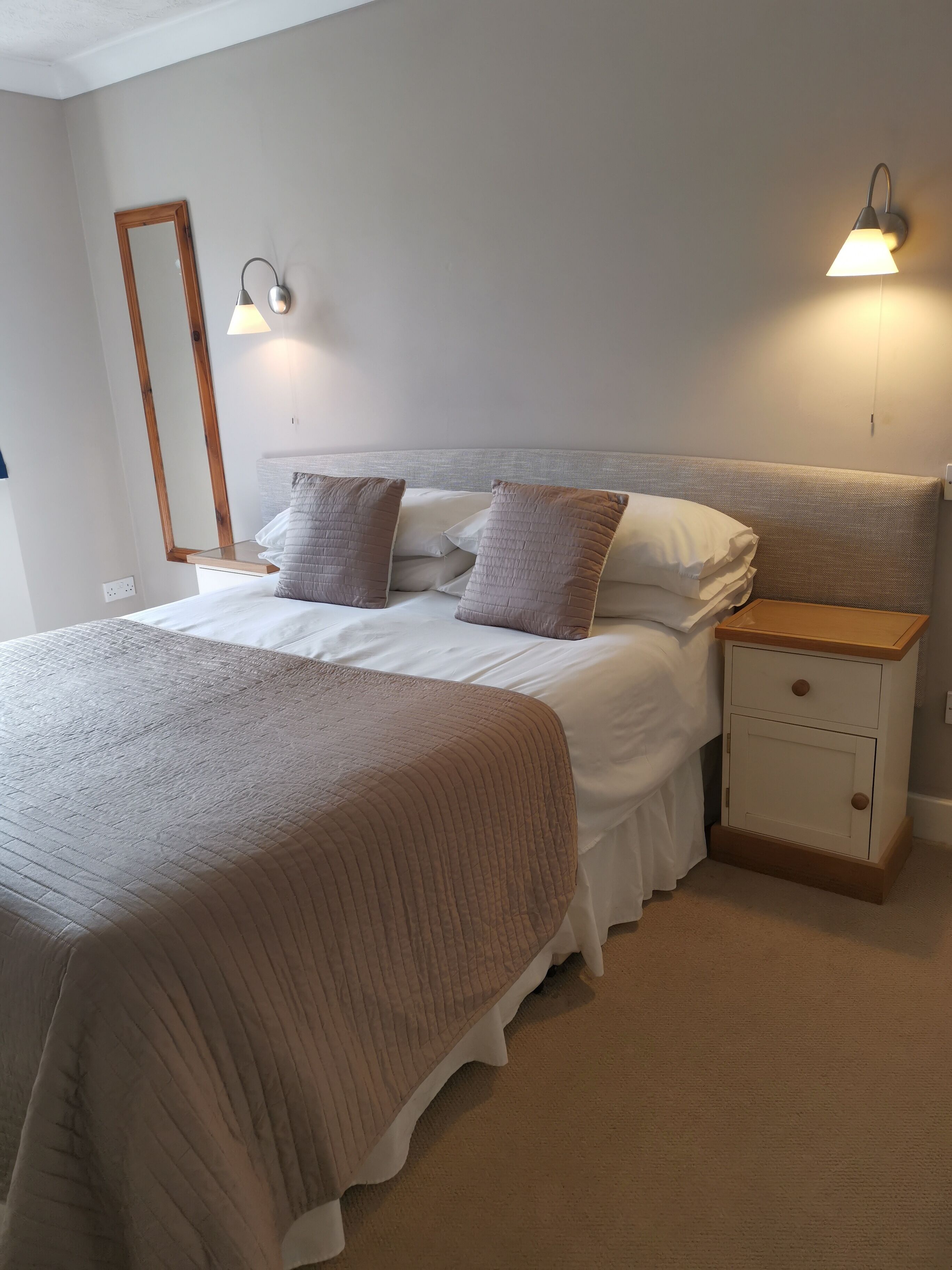 A neatly made bed with a brown quilt, a wooden nightstand with drawers, and two wall-mounted lamps.