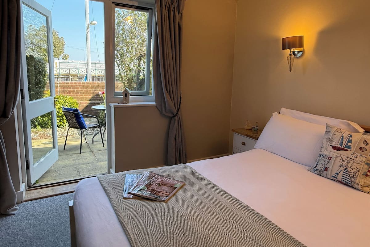 A hotel room with a bed, a window with curtains, a small table with a chair outside, and a wall-mounted lamp.