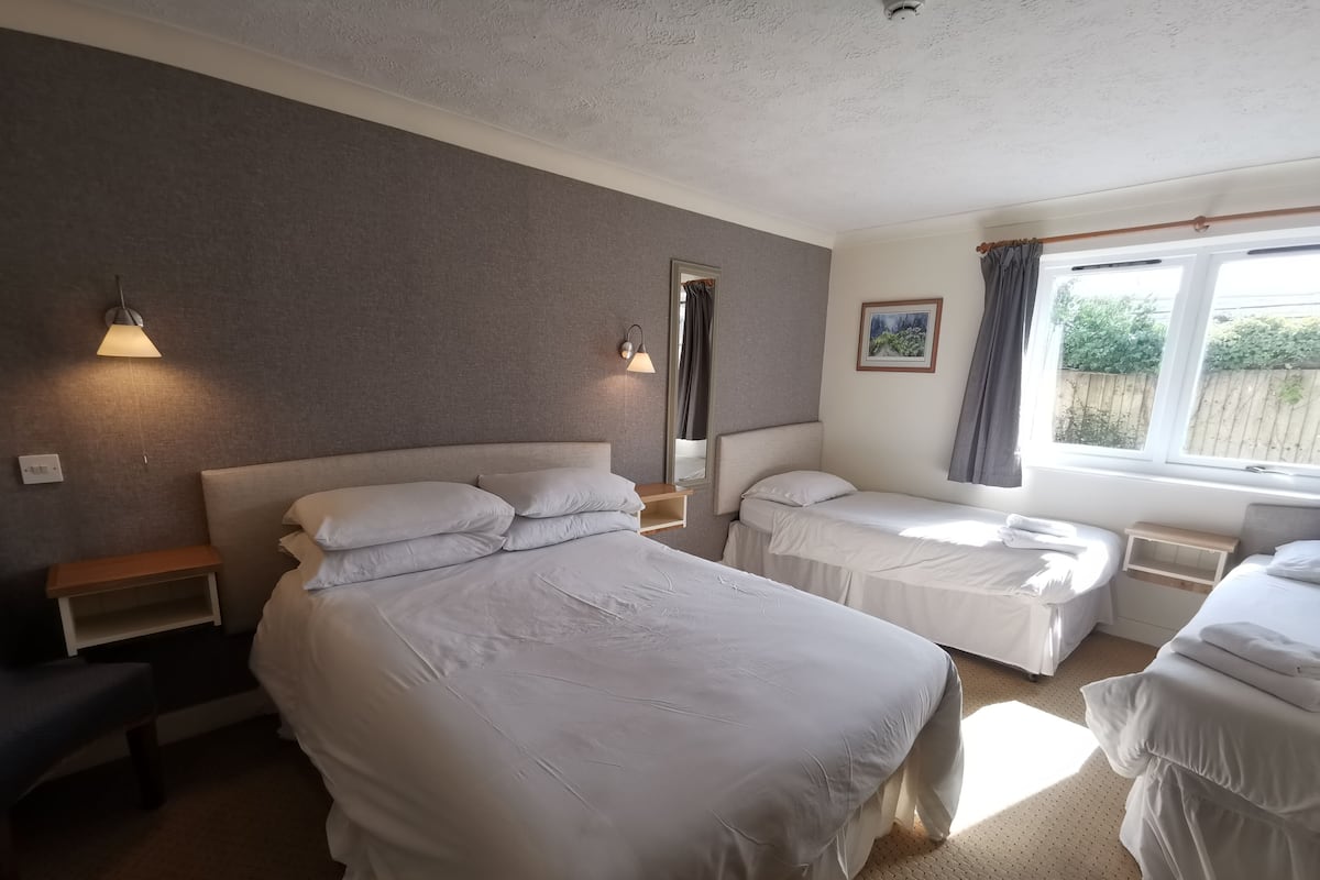 A hotel room with two beds, a bedside table, a nightstand, a window with curtains, and a wall-mounted light fixture.