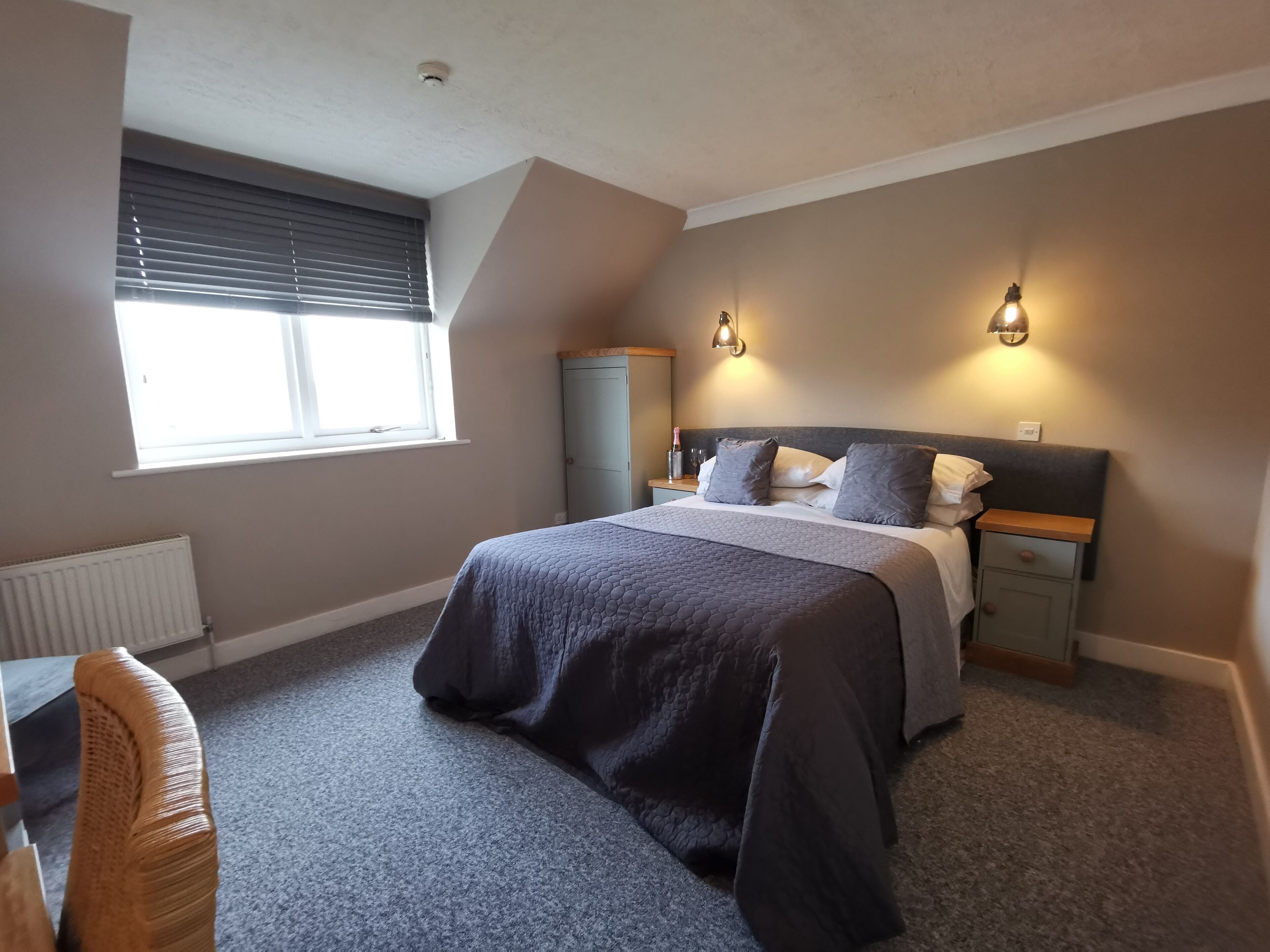 A bedroom with a bed, bedside tables, a wardrobe, a radiator, a window with blinds, and a chair.