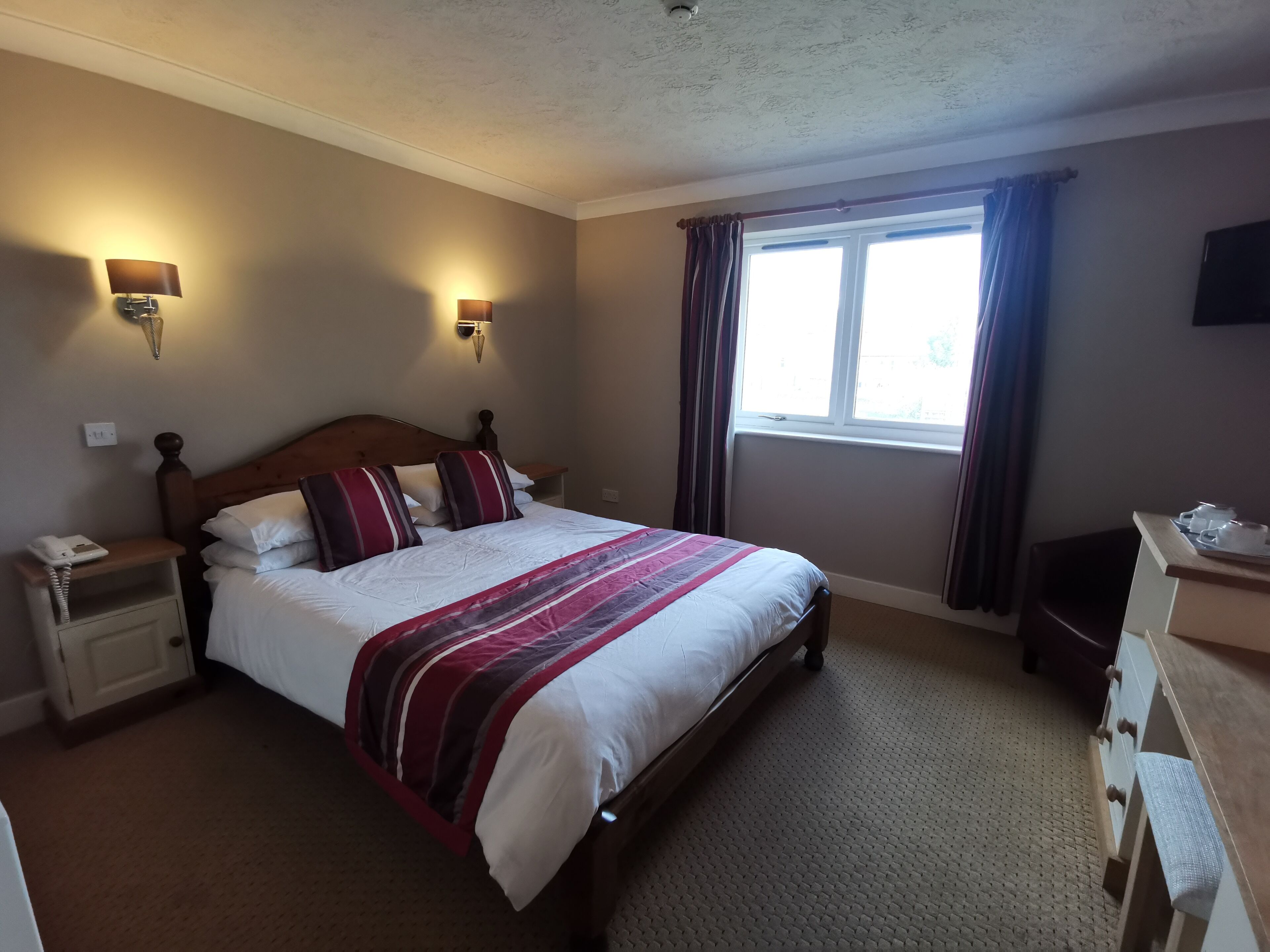 A neatly made bed with a striped blanket, two bedside lamps, a nightstand with a telephone, a window with curtains, and a dresser with drawers.