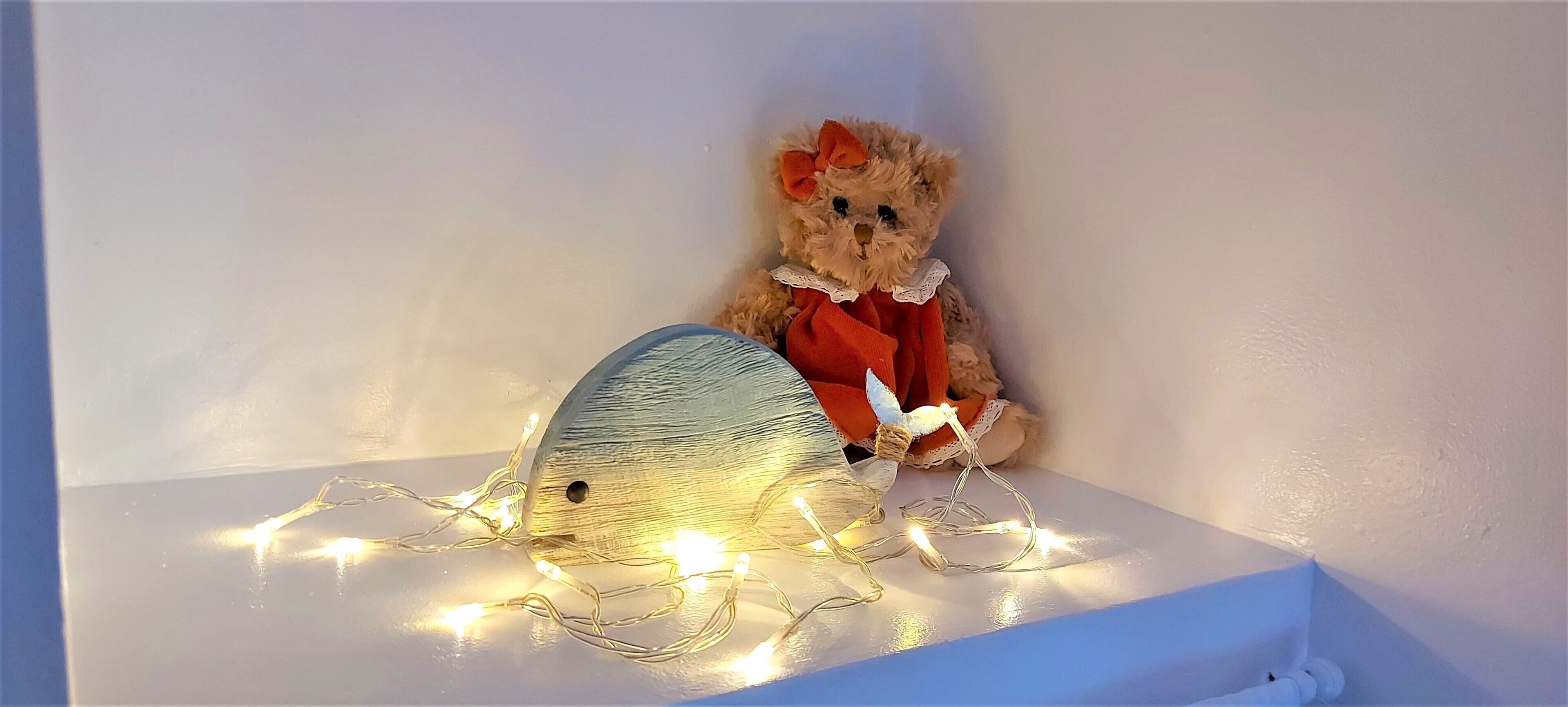 A teddy bear with a bow and a red bow tie sitting on a white shelf with string lights.