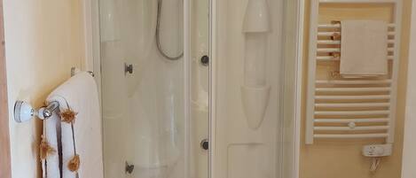 Un box de ducha con paredes de vidrio, una puerta blanca, un toallero con una toalla y un radiador blanco en la pared.