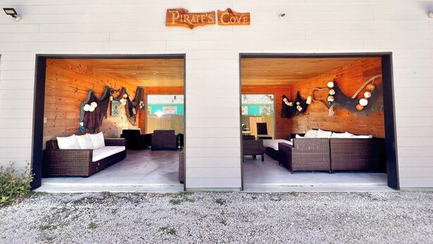 Una cabaña de madera con dos puertas grandes que dicen 'Pirate's Cove'. En su interior, hay sofás de mimbre y una mesa pequeña.