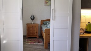 Ein Zimmer mit einer Schiebetür, die zu einem Schlafzimmer führt, einem kleinen Nachttisch, einem Bett, einem rot-grauen Teppich und einem weißen Tisch.