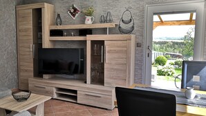 Ein modernes Wohnzimmer mit großem Fernseher, Holzschränken, einem Essbereich und Blick auf einen Garten.