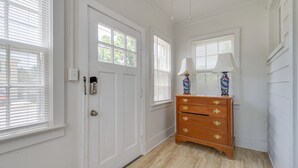 A hallway with white walls, a wooden floor, and a white door with a keyhole. There are two windows with blinds, a wooden dresser with a lamp, and a vase on top.