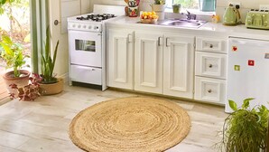 A kitchen with white cabinets, a stove, a microwave, and a refrigerator. There are potted plants and a woven rug on the floor. - One minute's walk to Freights Bay (Freights Bay)