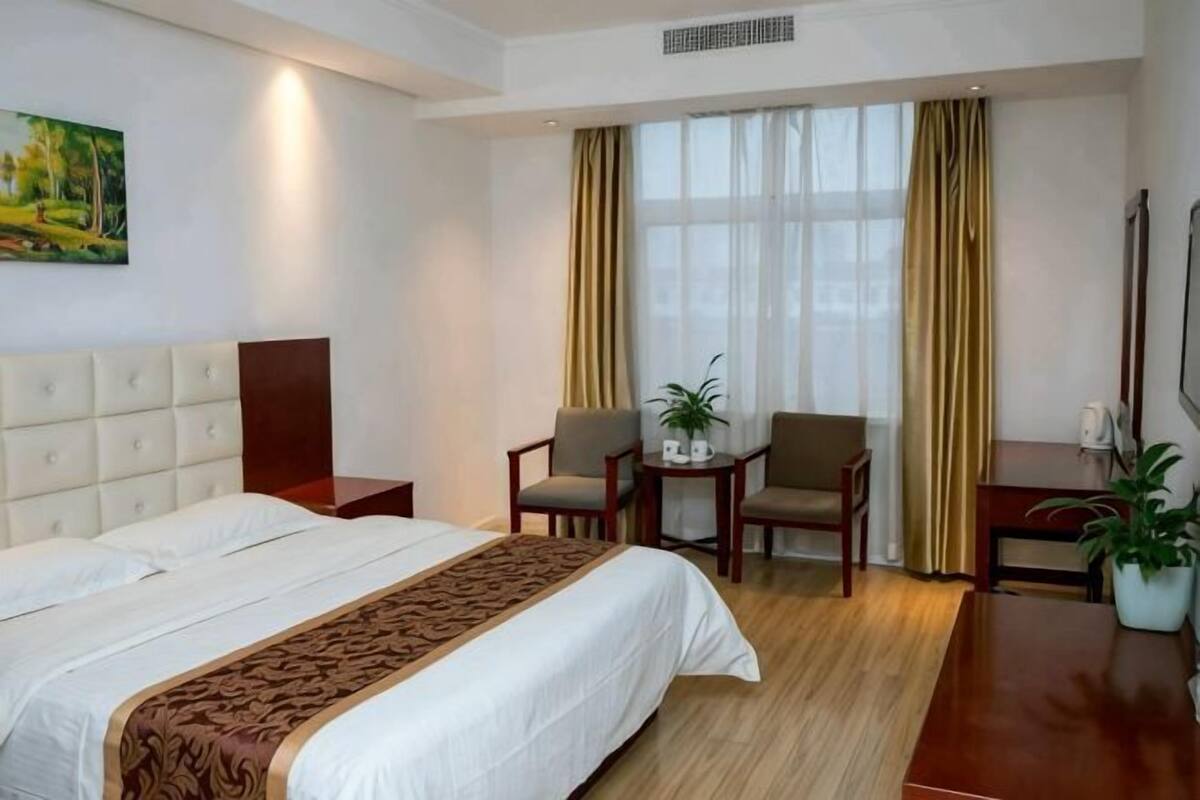 Ein Hotelzimmer mit einem großen Bett, einem Schreibtisch, zwei Stühlen, einem kleinen Tisch mit Pflanze und einem Fenster mit Vorhängen.