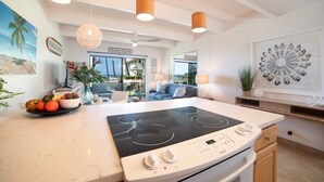 A modern kitchen with an induction cooktop, a dining area with a bowl of fruit, and a view of the beach. - Remodeled Condo Across Ocean w Pool AC-Shores of Maui 109 (Kihei)