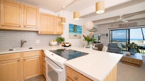 A modern kitchen with wooden cabinets, a built-in oven, and a large island with a stovetop. There is a dining area with a view of the ocean and a sign that reads "BEACH HOUSE". - Remodeled Condo Across Ocean w Pool AC-Shores of Maui 109 (Kihei)