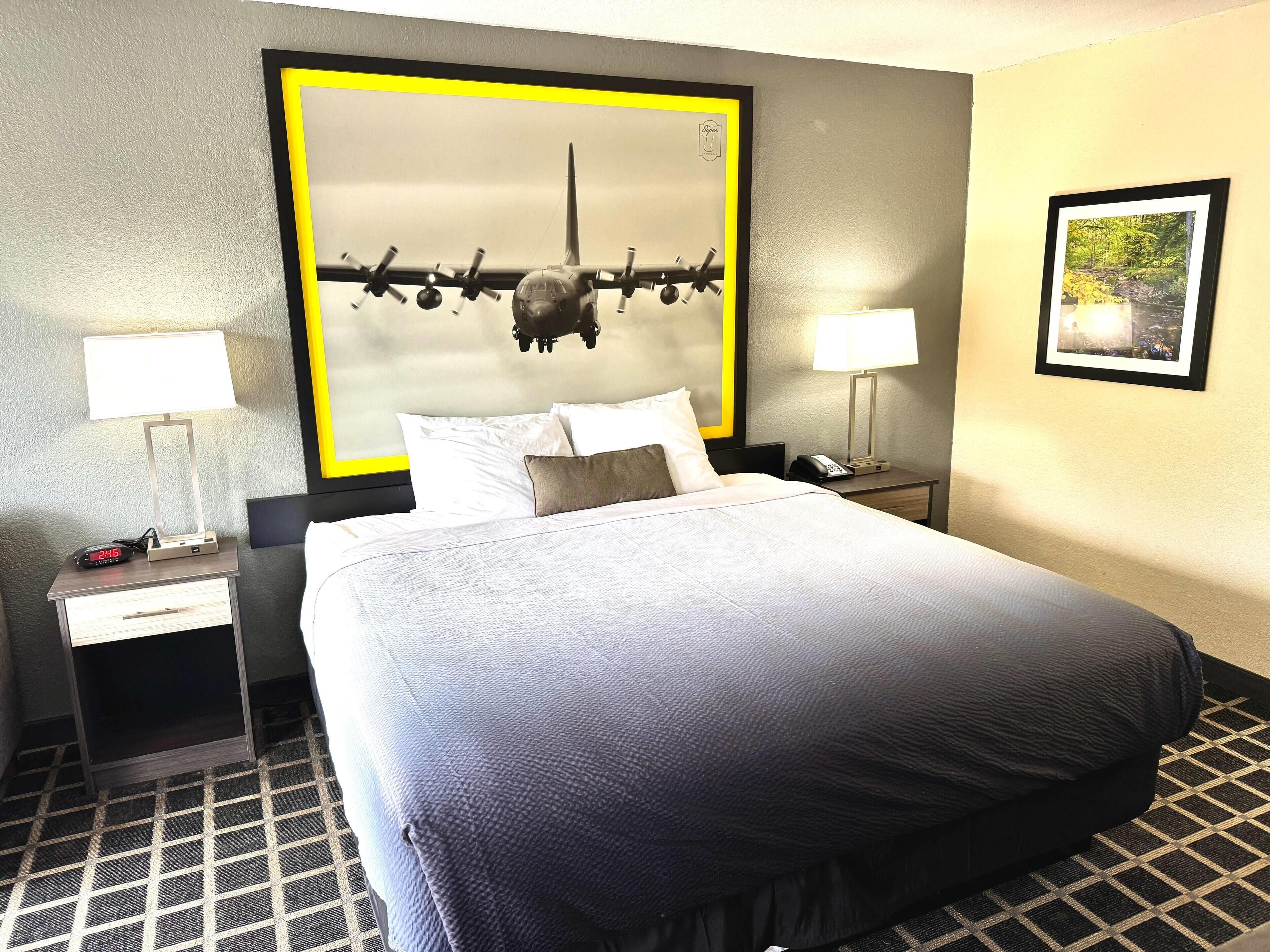 Una habitación de hotel con una cama grande, dos mesitas de noche, una lámpara de pared, un cuadro enmarcado y un reloj.