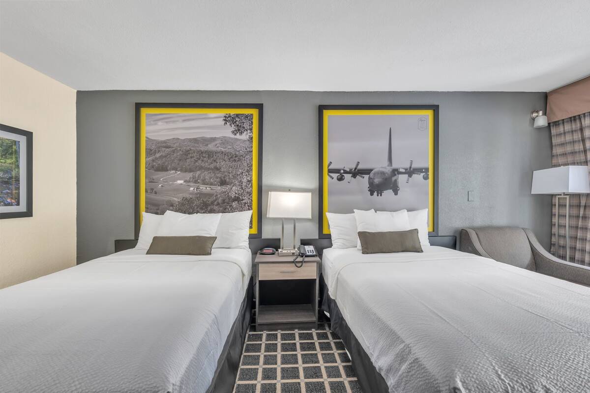 A hotel room with two beds, a nightstand with a lamp, a phone, and a view of mountains and a plane.