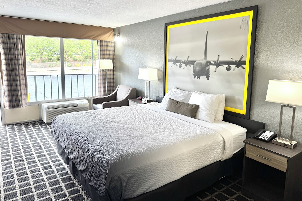 A hotel room with a large bed, a desk with a lamp, a chair, and a window with curtains.