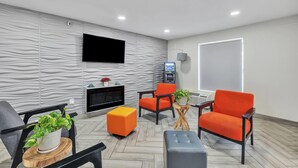 A modern lounge area with a flat-screen TV mounted on the wall, a coffee machine, and a seating arrangement including orange chairs and a grey ottoman. - Super 8 by Wyndham Morganton (Morganton)