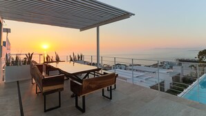 Une terrasse avec une table et des bancs, offrant une vue sur le paysage urbain et la mer au coucher du soleil.