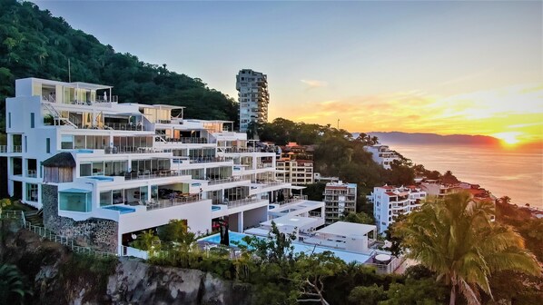 A coastal hotel with multiple balconies, a pool, and a view of the sunset over the ocean. - Breathtaking View with open design and Game Room (Puerto Vallarta)