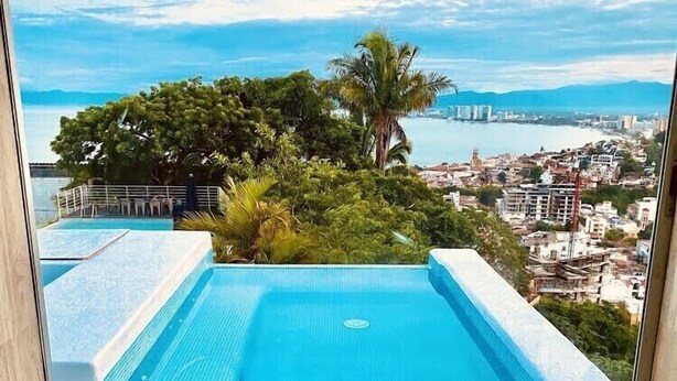 Une vue depuis une fenêtre sur une piscine, avec un paysage urbain en arrière-plan.