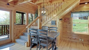 A wooden cabin interior with a dining table, chairs, a staircase, and a view of the outdoors.