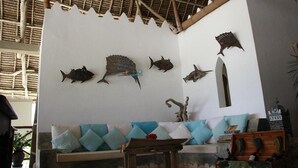 A room with a thatched roof, a white wall with fish wall art, a blue sofa with cushions, and a wooden chest. - Mvuvi Boutique Resort (Kiwengwa)