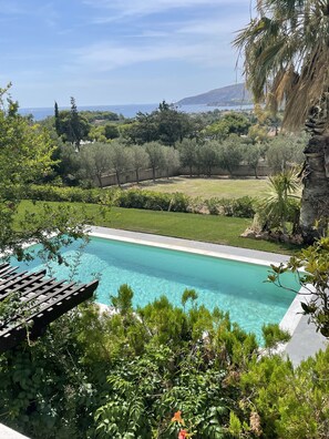 Una piscina amb aigua transparent de color blau, envoltada de vegetació exuberant i vista al mar i a les colines al fons.