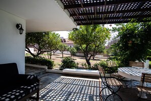 An outdoor patio with a round table and benches, surrounded by trees and a view of buildings in the background. - Villa Sounio (ATTICA)