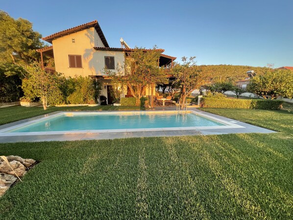 A swimming pool surrounded by well-maintained grass, a house with a tiled roof, and a clear sky. - Villa Sounio (ATTICA)