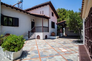 A courtyard with a tiled floor, a white building with a brown roof, and a large planter with green plants. - Villa Sounio (ATTICA)