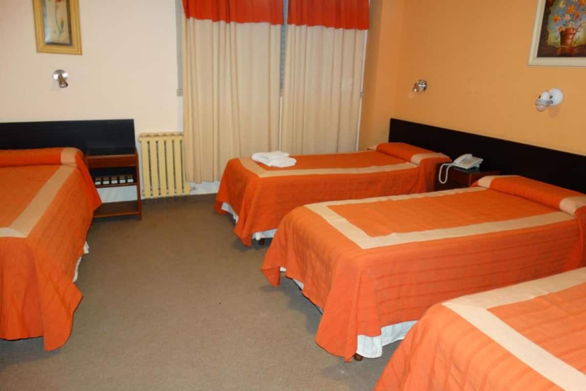 Quarto de hotel com três camas, paredes bege e uma janela com cortinas laranja.